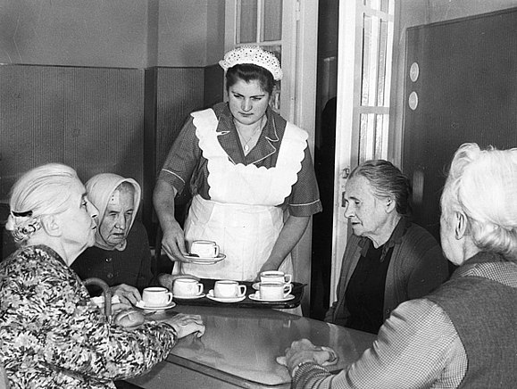 Servierkraft mit weißer Schürze und Häubchen beim Anbieten von Kaffee mit vier weiblichen Dauergästen im Speisesaal 1961