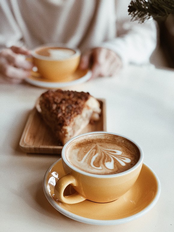 Symbolbild Kaffeetassen und Kuchen auf einem Tisch in Nahaufnahme. Im Hintergrund Frauenhände, sitzend, eine Kaffeetasse in der Hand.